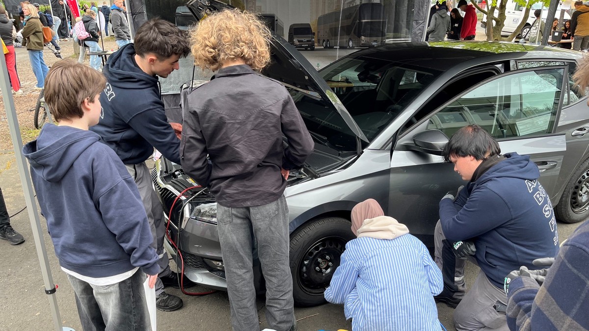 Mehrere Schüler in dunklen Hoodies besichtigen gemeinsam die geöffnete Motorhaube eines silbernen Autos bei einer Handwerkspräsentation.