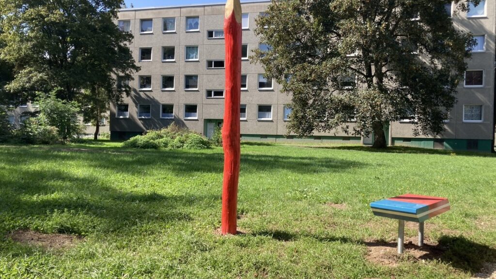 Grüne Wiese mit roter Stange und farbigen Spielplatten vor mehrstöckigen Wohngebäuden.